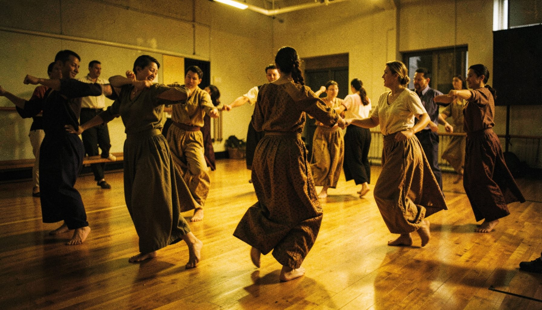 A warm, dimly lit dance studio with hardwood floors, a group of dancers moving in unison in loose traditional attire, deep browns and golds dominating the scene, candid and atmospheric