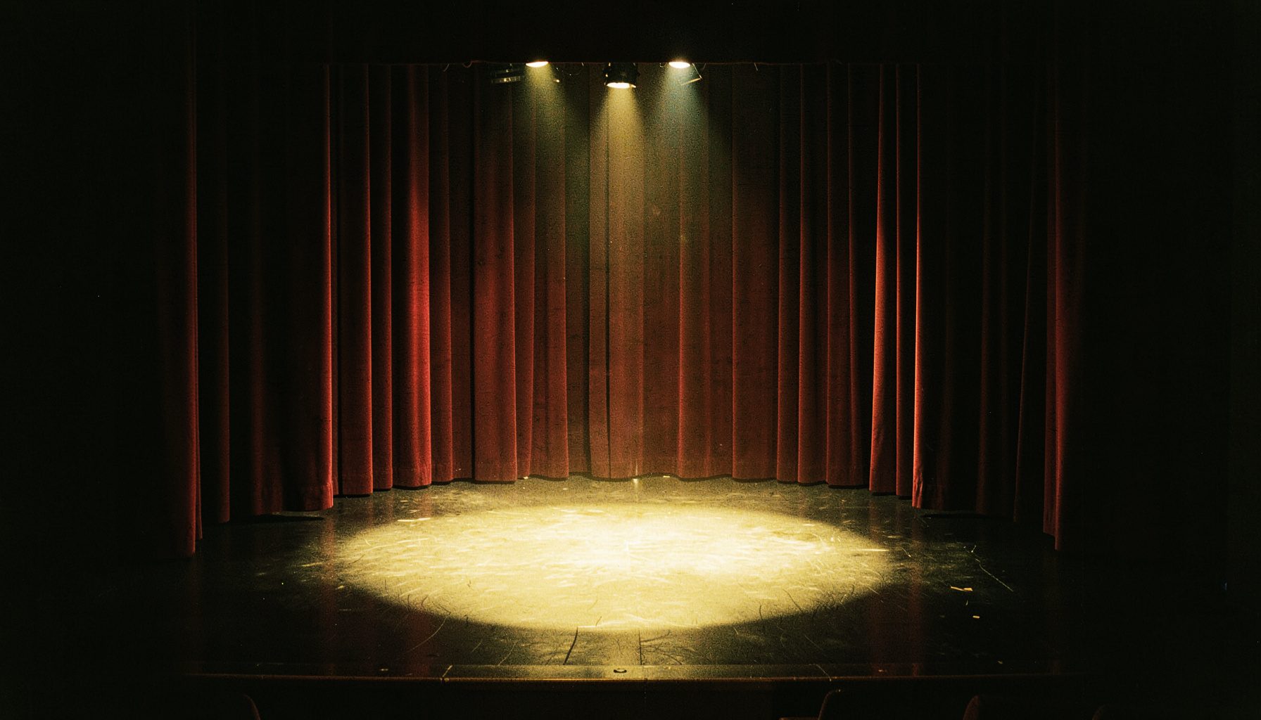 A theater stage viewed from the wings, deep burgundy curtains framing a spotlight pool on the floor, rich shadows and warm highlights