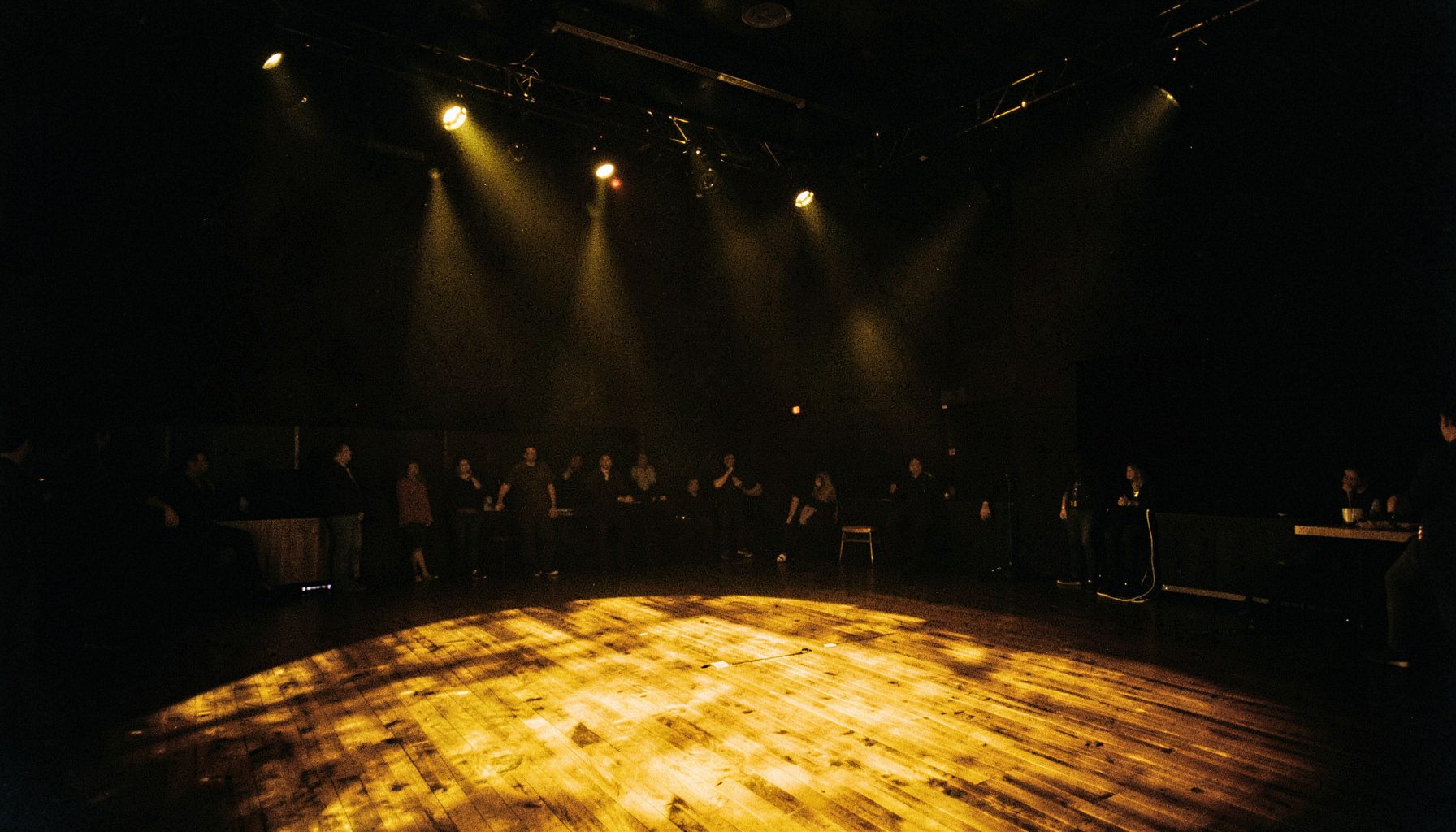 A dimly lit performance space with warm amber stage lights casting dramatic shadows across a wooden floor, deep blacks and rich golds dominating the scene
