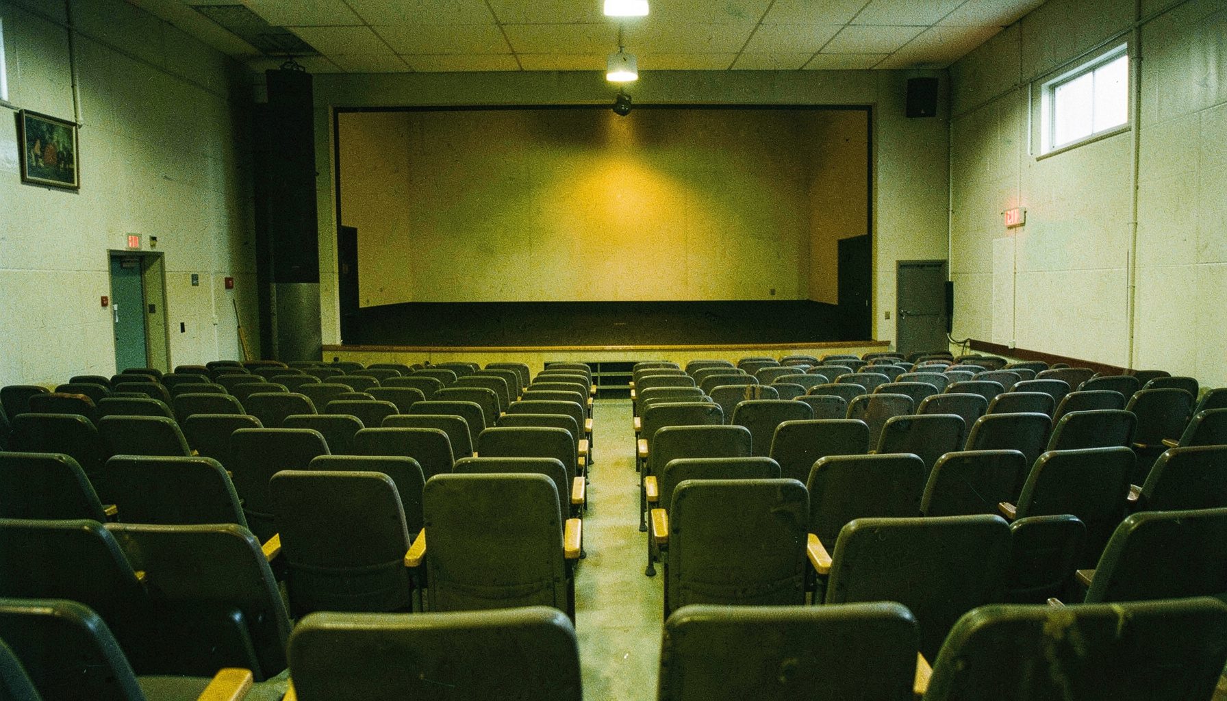 A high school auditorium with rows of empty seats and a bare stage, cool institutional lighting with warm exit-sign glow