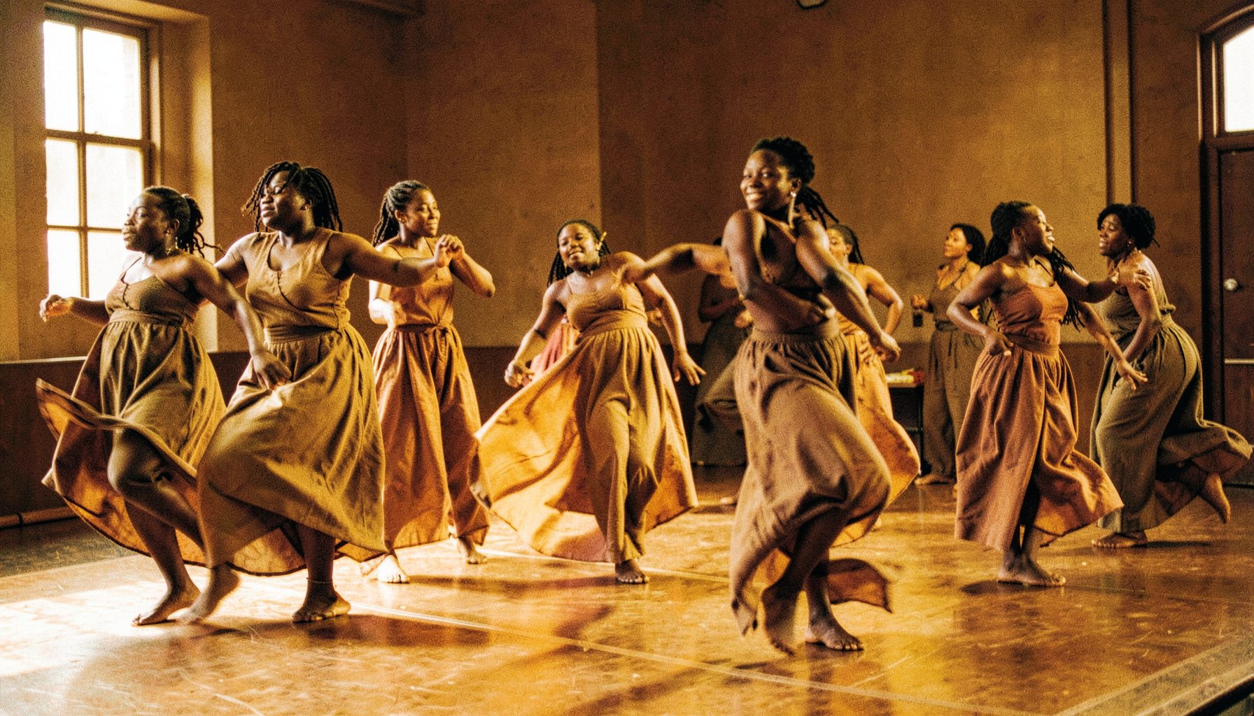 A group of Black women dancers in motion, wearing flowing earth-toned fabrics, moving together in a warm-lit studio space with deep amber and brown tones.