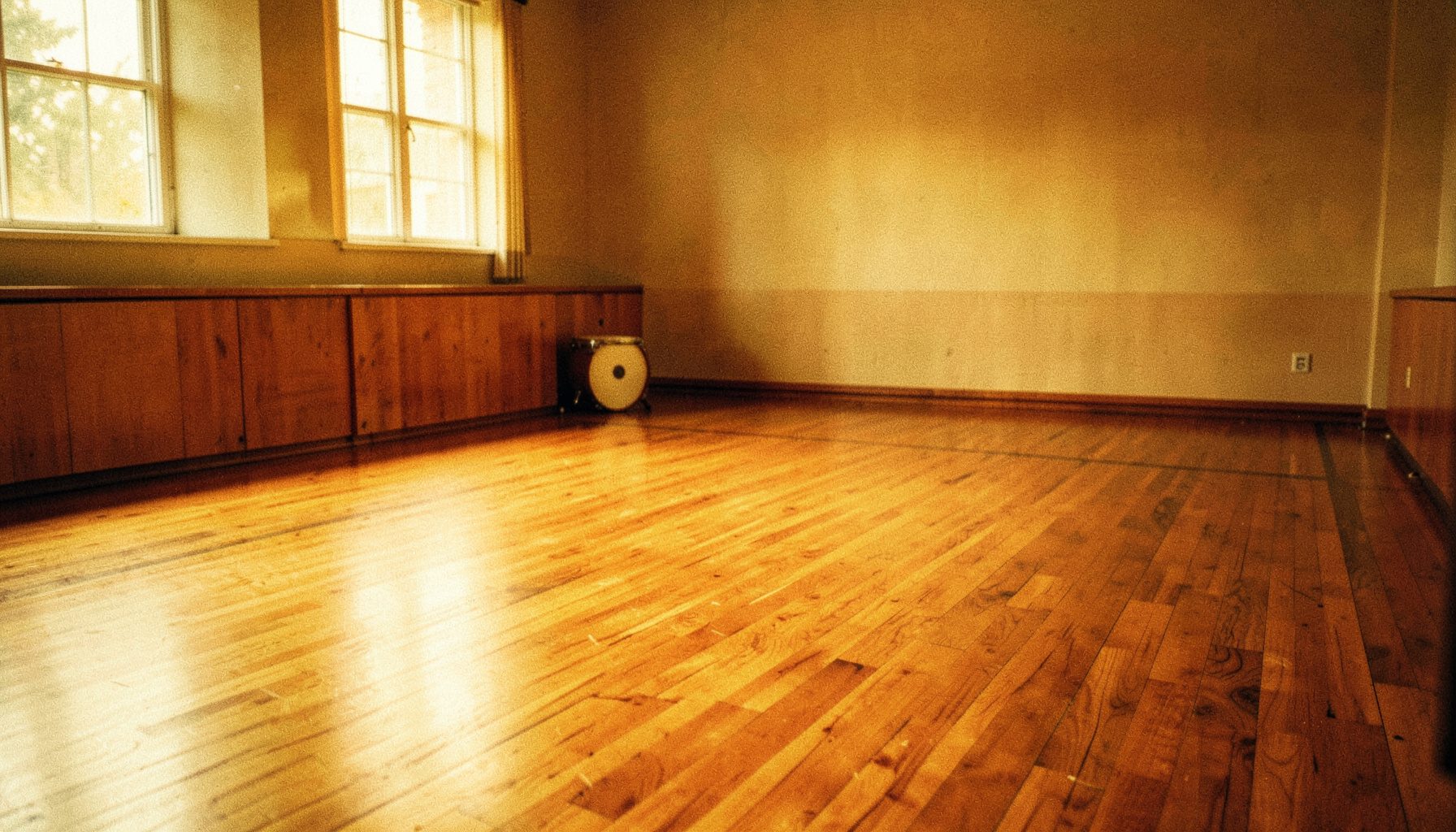 A warmly lit dance studio with polished wooden floors, soft amber and brown tones filling the space, a single drum resting in the corner suggesting quiet anticipation.