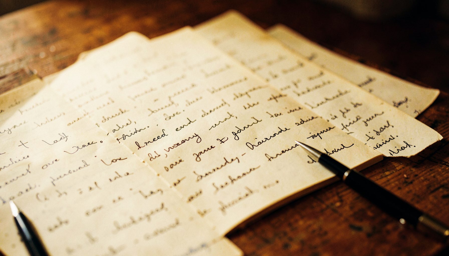 A softly lit close-up of handwritten journal pages spread across a wooden surface, warm amber and deep brown tones, with a pen resting beside the paper.