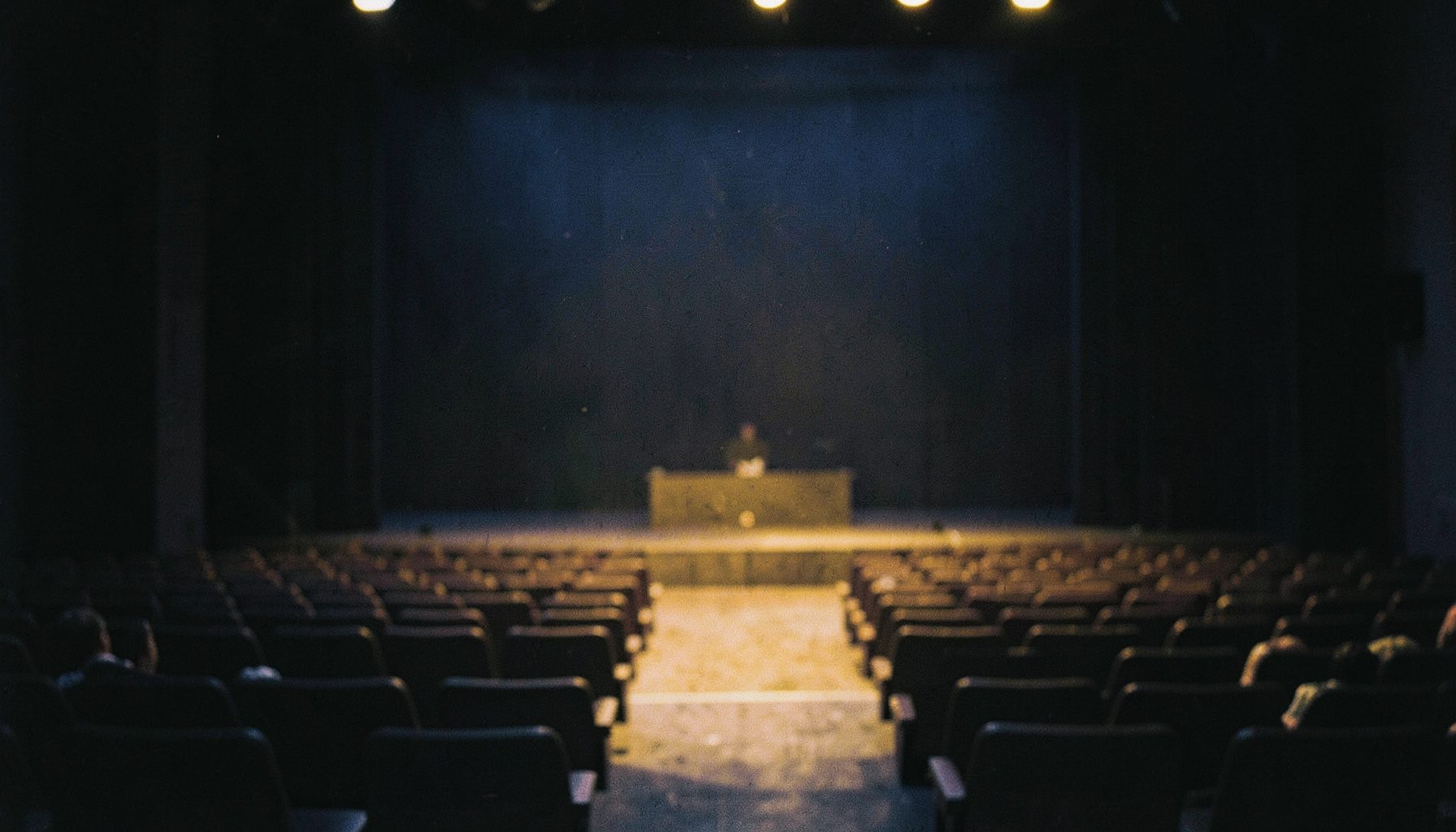 A dreamy, soft-focus scene of a stage at twilight, deep indigo and muted gold, with empty seats stretching into shadow.
