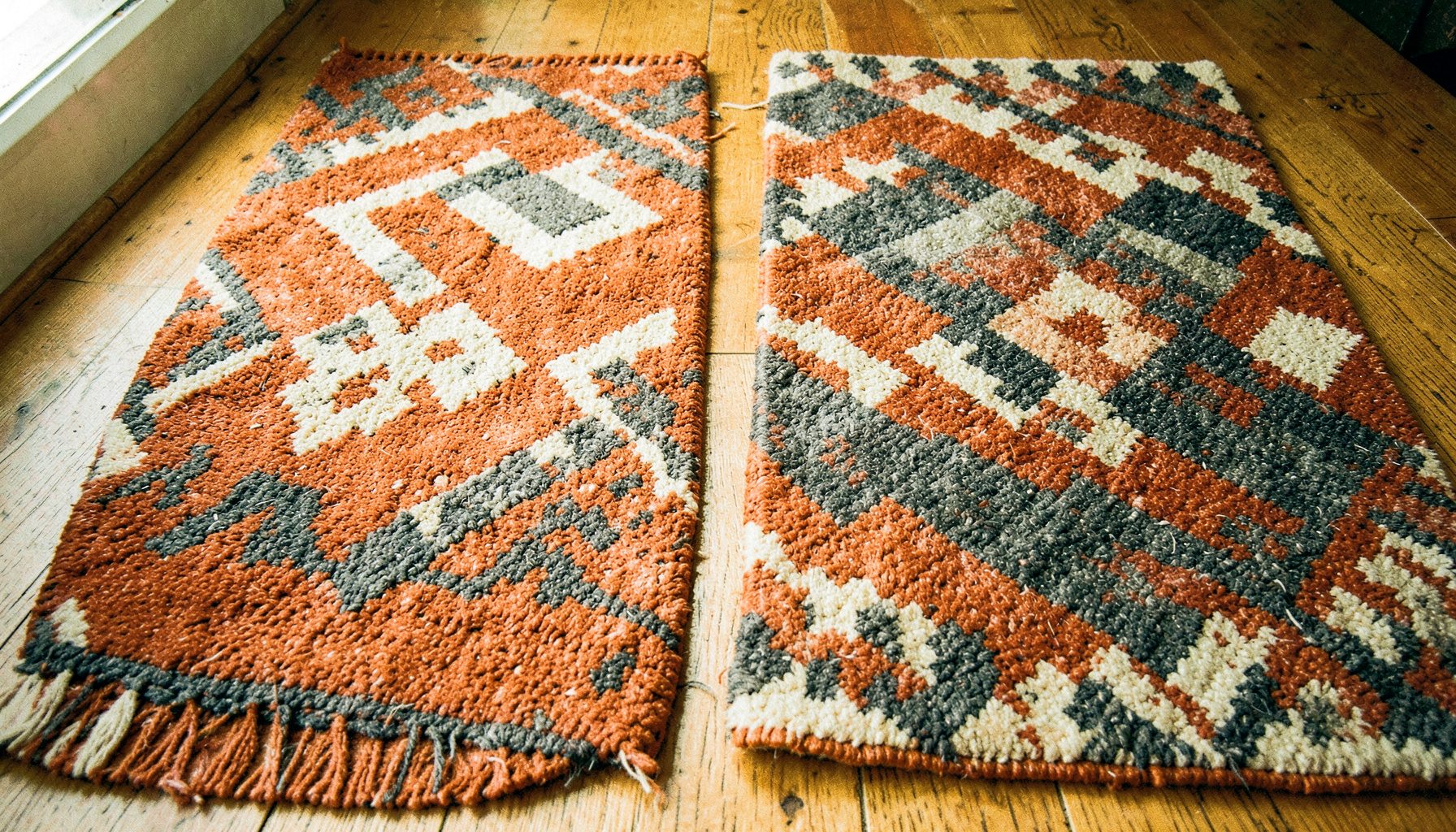 Two woven rugs with bold geometric patterns in rust, cream, and charcoal, photographed in natural light on a wooden floor.