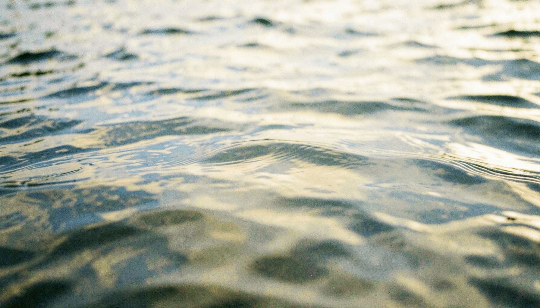 A close view of rippling water reflecting overcast sky, muted silver and slate blue, with subtle tension at the surface.
