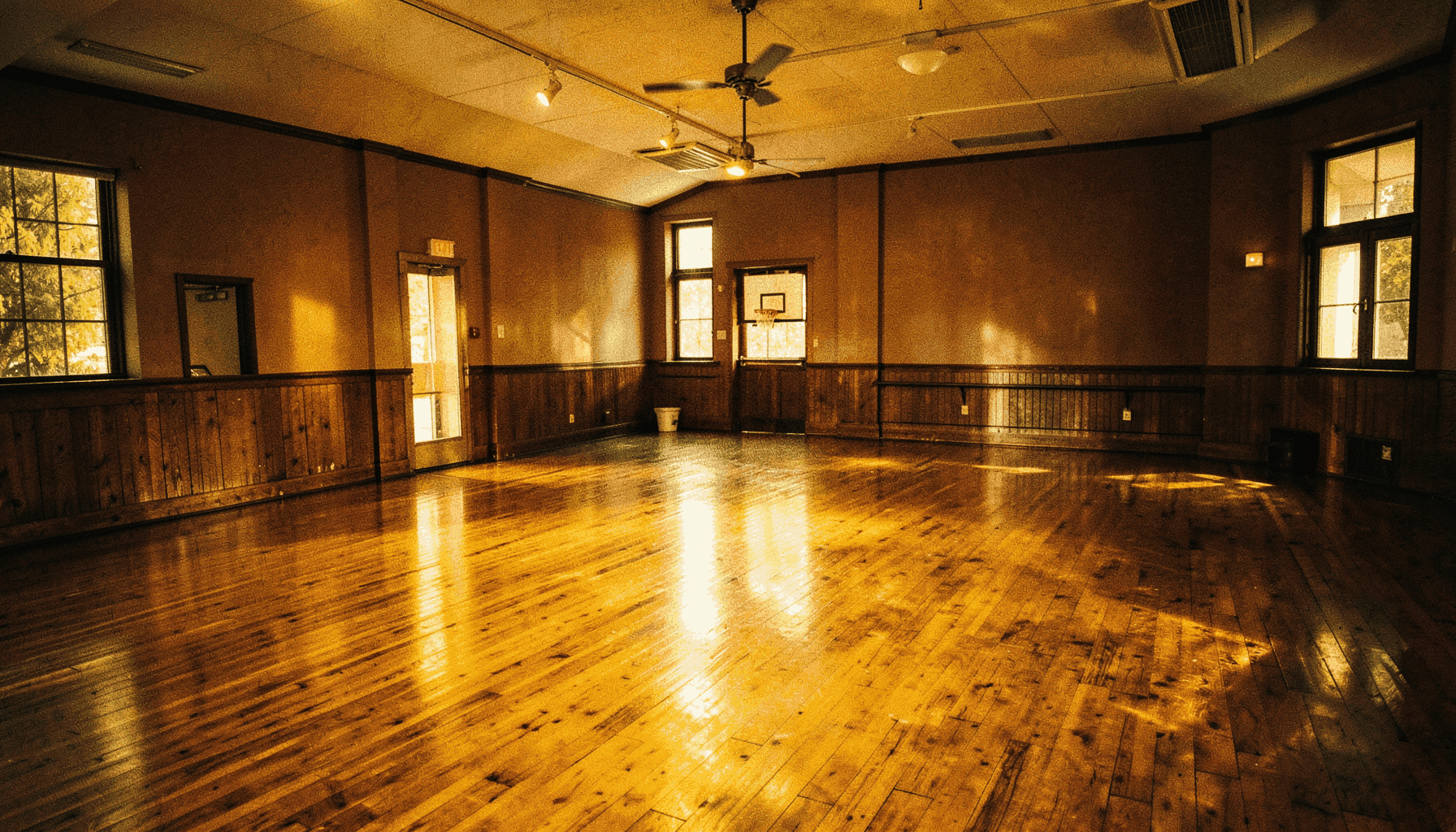A warmly lit community dance studio interior with rich amber and deep brown tones, wooden floor reflecting soft light, empty and ready for gathering.
